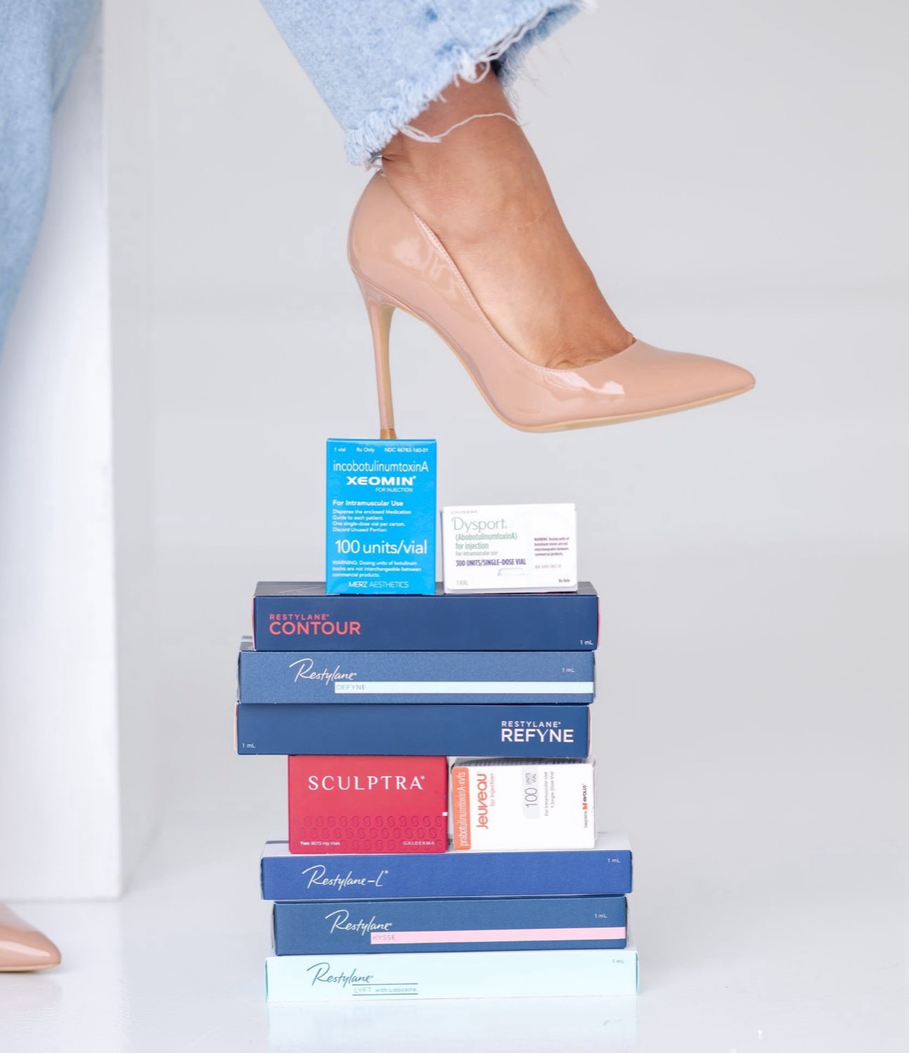 A stack of injectable cartons — Xeomin, Dysport, Restylane, Sculptra and Jeuveau — with a nude heel resting on top.